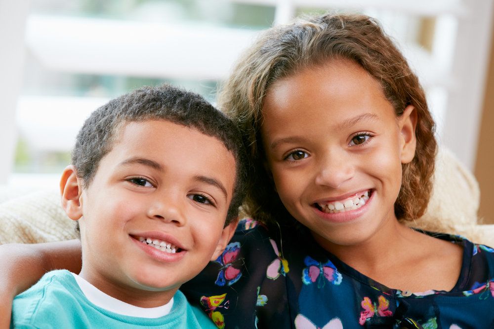 A young brother and sister with beautiful smiles sit on the couch smiling together.